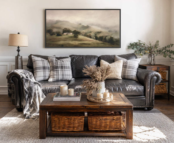 This horizontal canvas wall art features a misty, atmospheric landscape of rolling hills with a small house, rendered in a soft, muted palette and a painterly style, displayed on a.