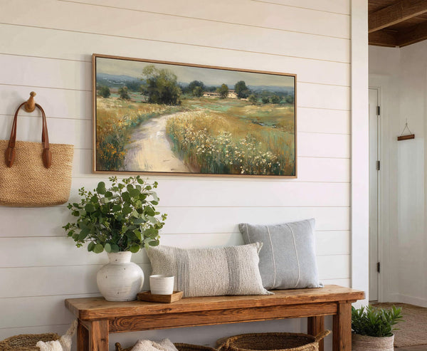 This canvas wall art, featuring a landscape with a dirt road winding through a field of wildflowers and a distant house, is framed in natural wood. It hangs above a.