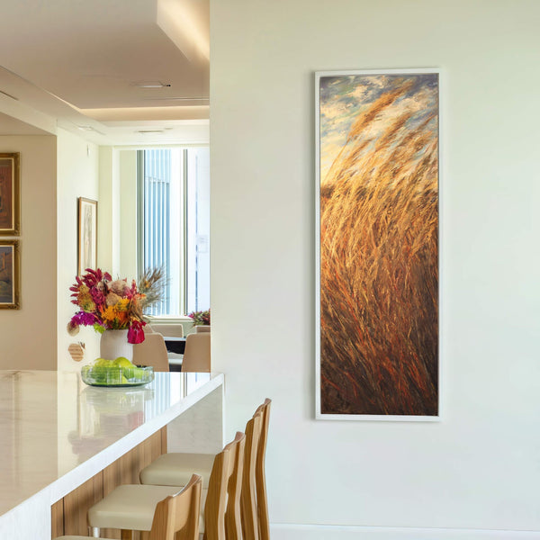This vertical canvas wall art features tall grasses in warm, earthy tones. It is framed in white and displayed on a plain white wall, with wooden bar stools visible in.