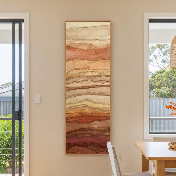 This vertical canvas print features an abstract landscape with warm earthy tones and organic layered shapes, framed in natural wood and displayed in a room with a light beige wall.