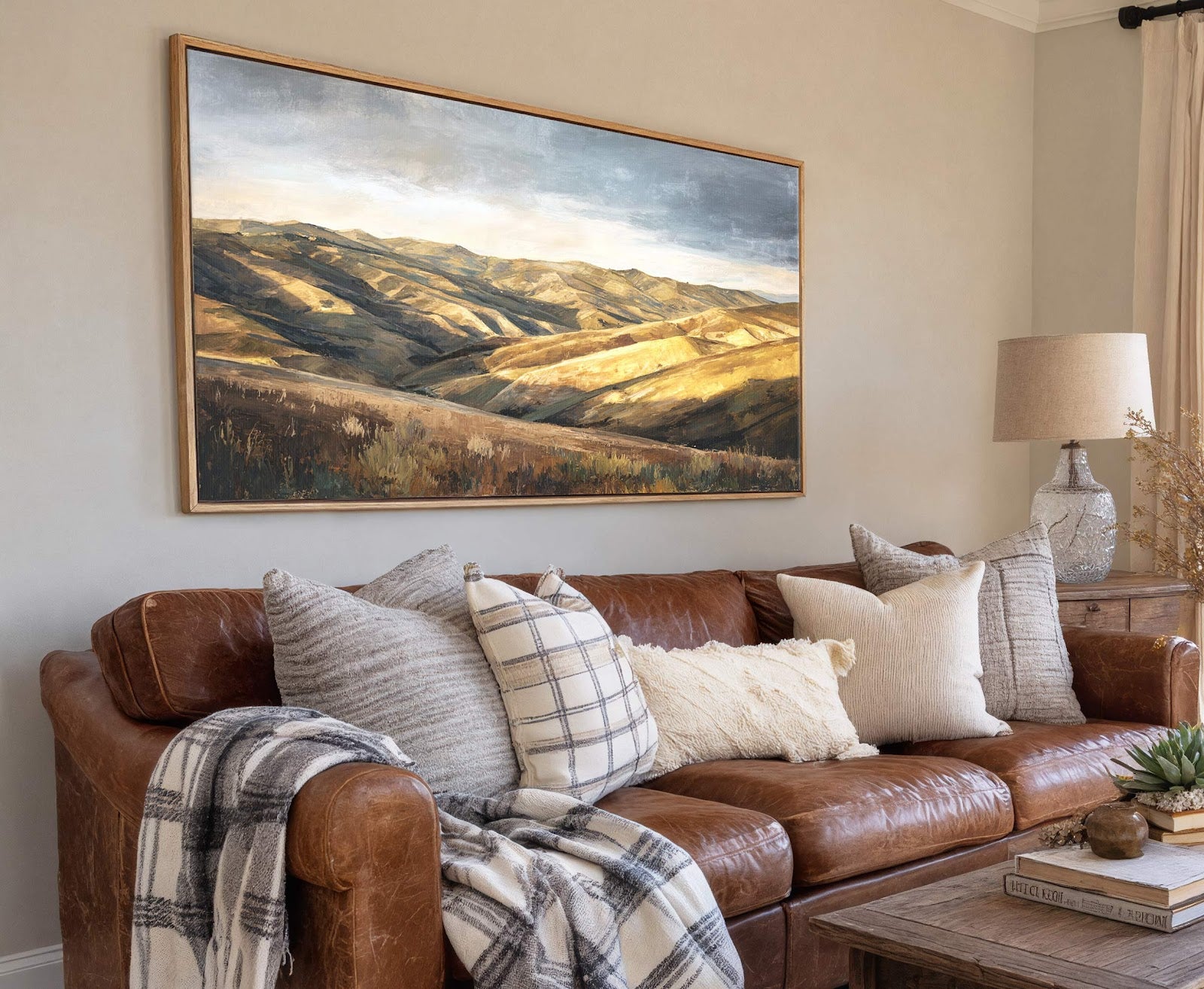 A horizontal canvas wall art print depicting rolling hills in a muted palette of greens, browns, and blues, framed in natural wood, hangs above a brown leather sofa with decorative.