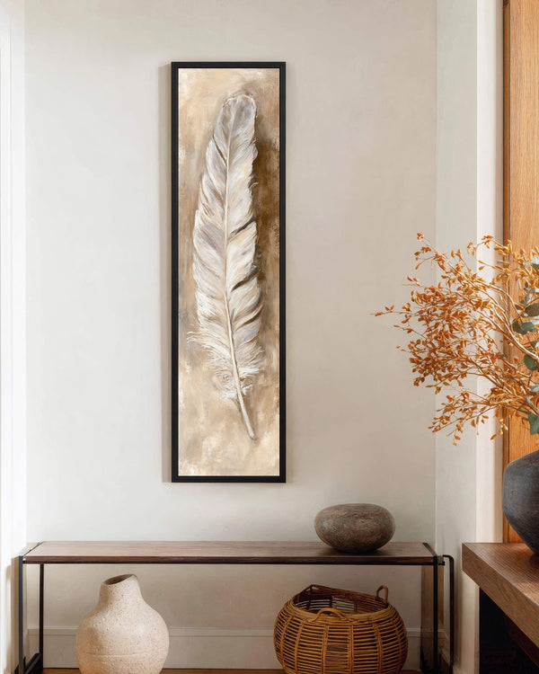 This vertical art print features a textured white feather in a neutral tan palette, shown in a black floater frame on a cream wall above a dark wooden console.