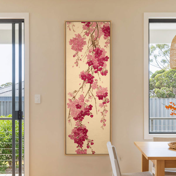 This vertical, framed canvas wall art depicts branches with pink and magenta cherry blossoms in a traditional style against a muted gold background, displayed on a cream-colored wall near a.