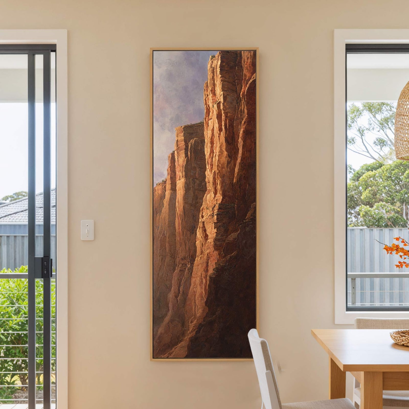 A vertical canvas wall art print depicts majestic rock formations with a warm, earthy palette and realistic style, displayed on a beige wall next to a window.