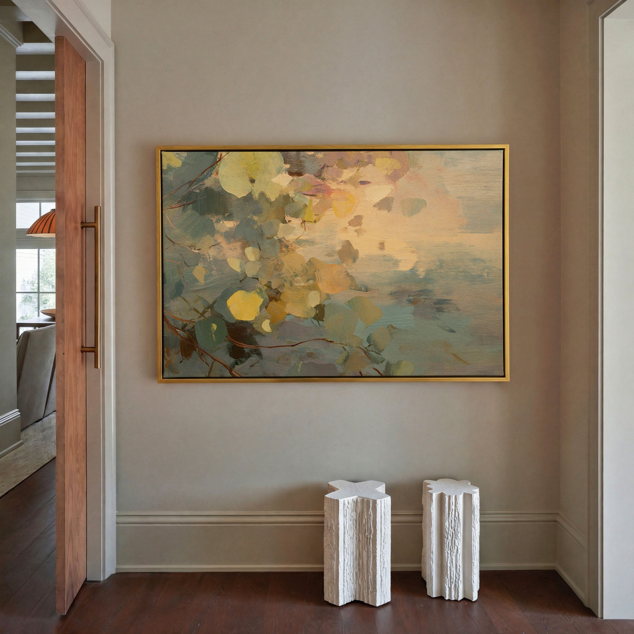 Abstract botanical canvas print in gold floater frame hanging in a warm taupe entryway — featuring loose leaf forms in sage green, amber, and soft gold above a misty teal background, styled above two white sculptural side tables with dark hardwood floors