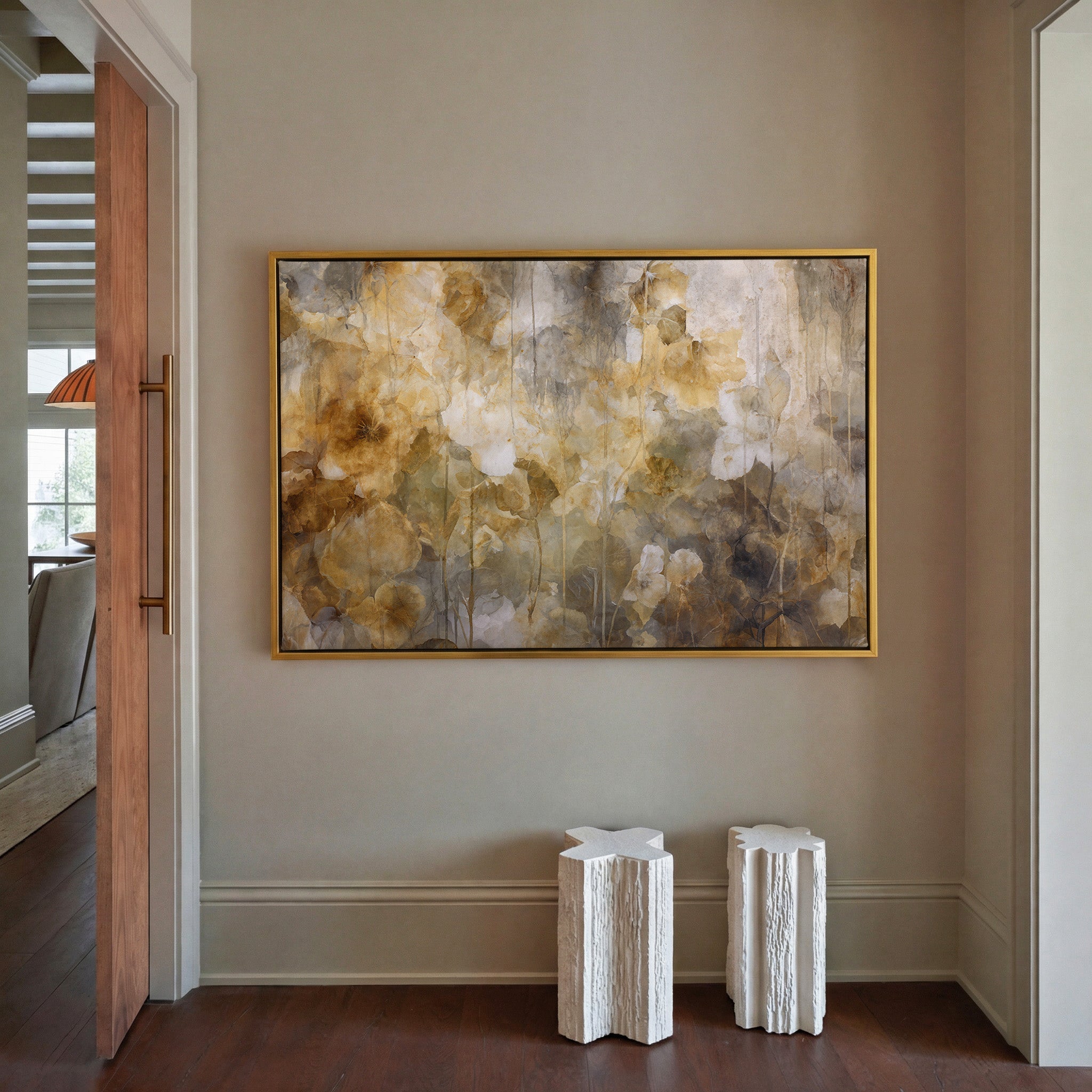 Moody abstract botanical canvas print in gold floater frame hung in a warm taupe entryway — layered floral and leaf forms in amber, warm brown, charcoal, and cream with delicate vertical drip lines, styled above two white sculptural side tables on dark hardwood floors