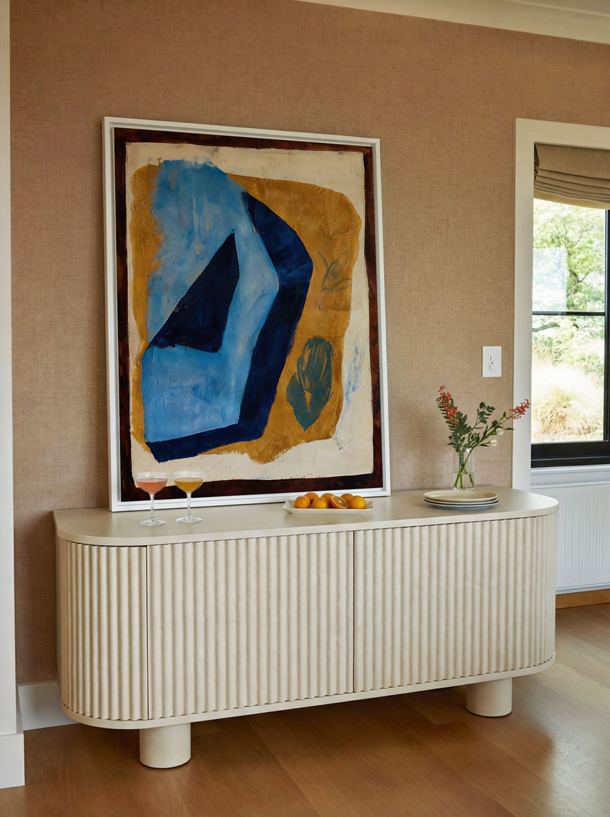 Bold abstract geometric canvas print in white floater frame above a cream fluted sideboard — large angular cerulean and deep navy form against a raw ochre ground with dark umber edges and faint botanical sketches, styled in a warm linen-walled dining space