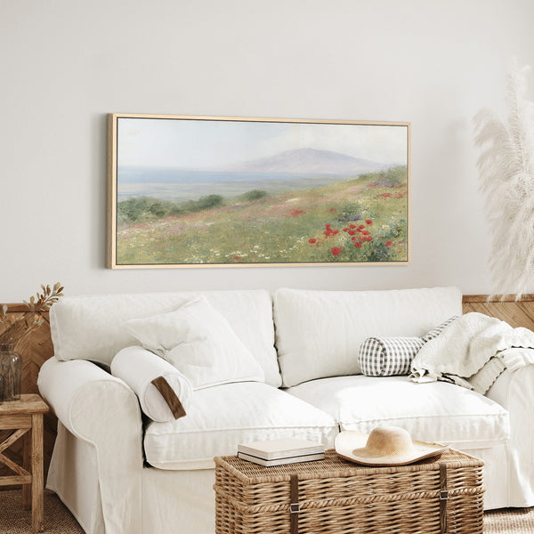 Large 30x60" natural framed impressionistic landscape art print depicting a vibrant wildflower meadow with red poppies in the foreground, leading to hills, a calm sea, and a hazy mountain under a soft sky, displayed above a white slipcovered sofa in a bright, relaxed living room.