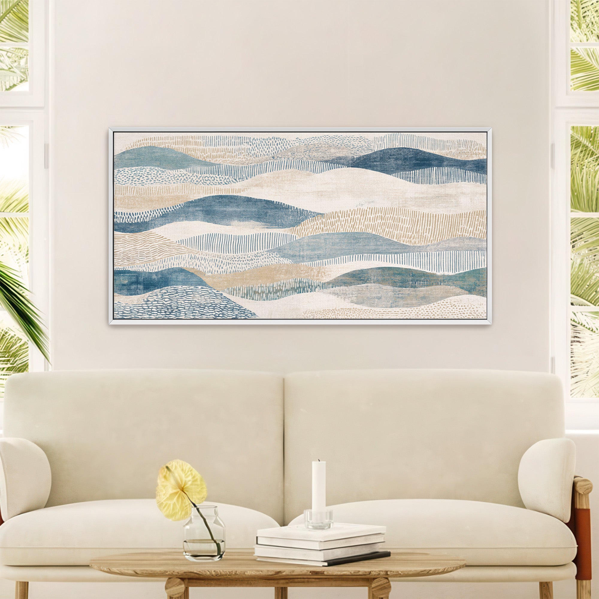 White-framed abstract art print featuring layered, flowing shapes with fine textured lines and patterns in muted blues, beiges, and whites, resembling rolling dunes or waves, displayed above a cream sofa in a bright, modern living room with large windows