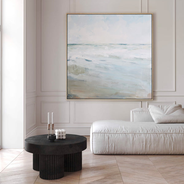 "Large natural wood-framed abstract seascape painting featuring soft, textured waves in blues, whites, and creams under a hazy sky, displayed above a white modular sofa in a modern living room with neutral walls