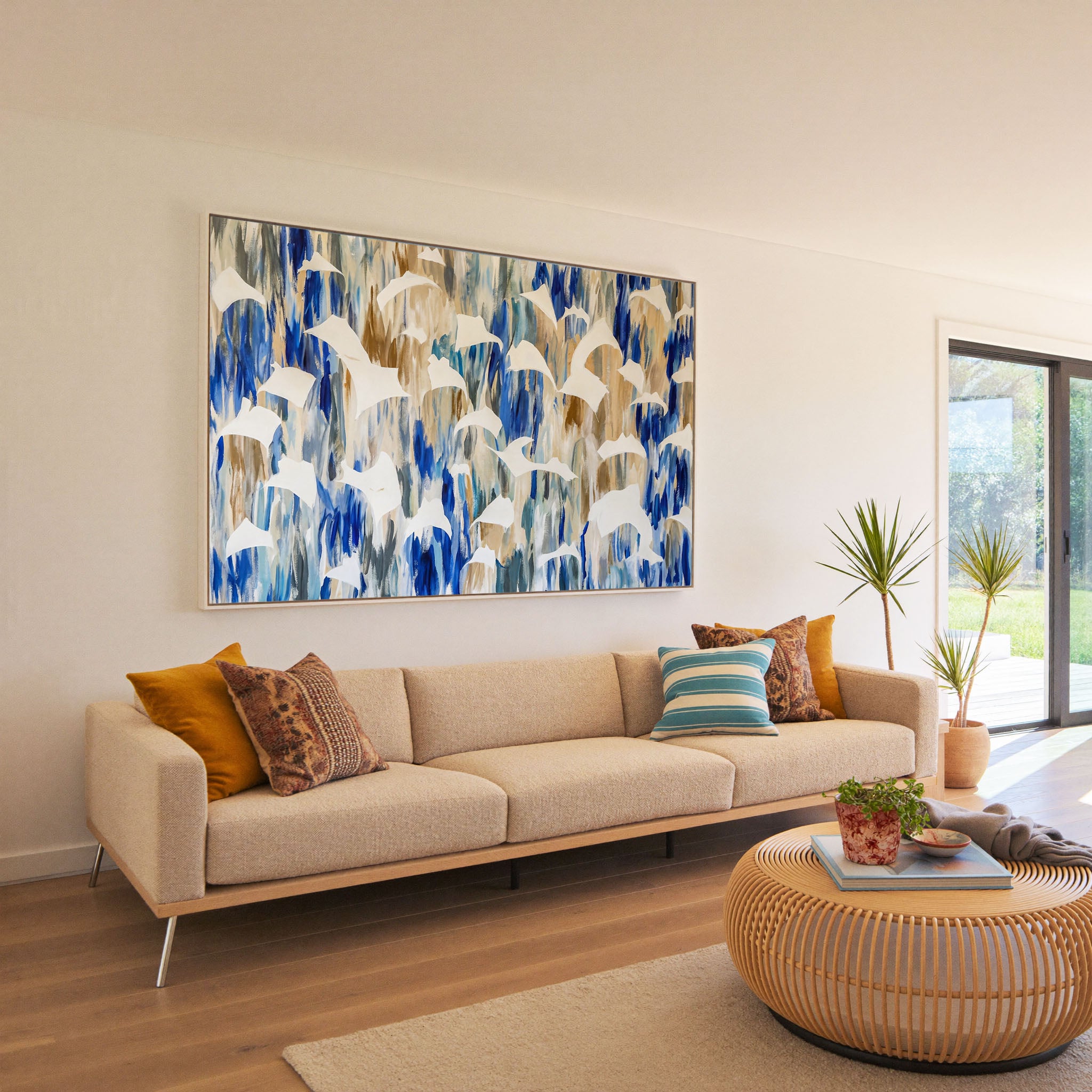 Large abstract canvas print in natural floater frame above a cream sofa — white bird silhouettes in flight against a vibrant cobalt blue and warm sandy gold abstract background, styled in a bright contemporary living room with rattan coffee table and mustard throw pillows