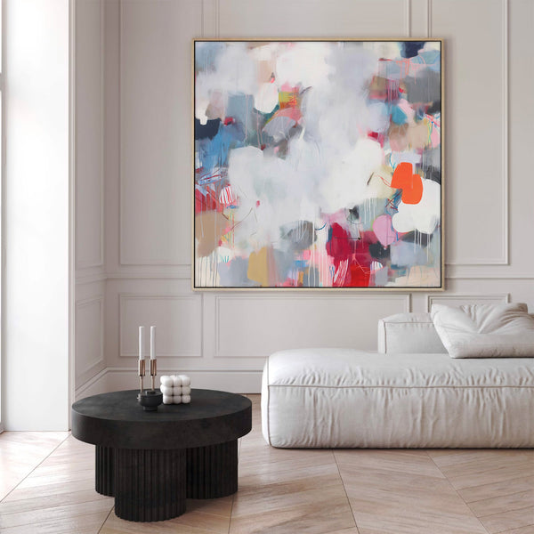 Large natural framed abstract painting featuring dynamic, layered forms in shades of white, gray, blue, red, and orange, displayed above a white modular sofa and fluted black coffee table in a modern living room with neutral walls