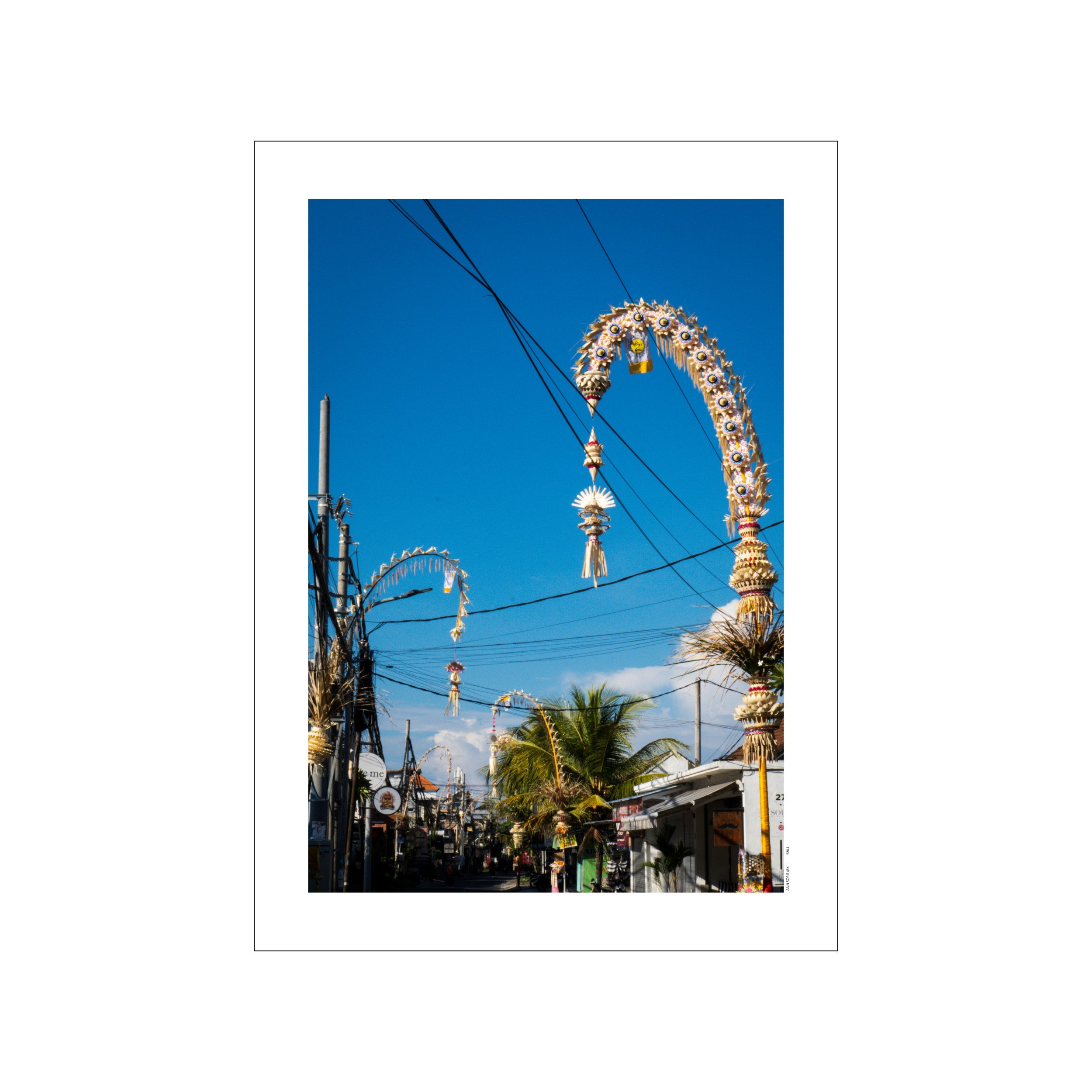A street scene in Canggu, Bali, with elaborate, woven Galungan decorations hanging overhead against a bright blue sky, framed by palm fronds and buildings. The artwork captures a festive, cultural atmosphere with warm, natural tones.