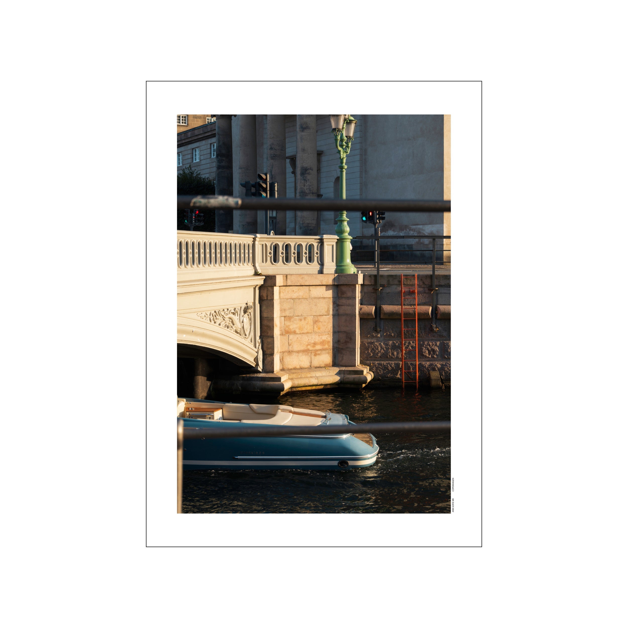 A tranquil Copenhagen canal scene at golden hour, with a modern blue boat passing under an ornate bridge, illuminated by warm sunlight and a vintage green lamppost.
