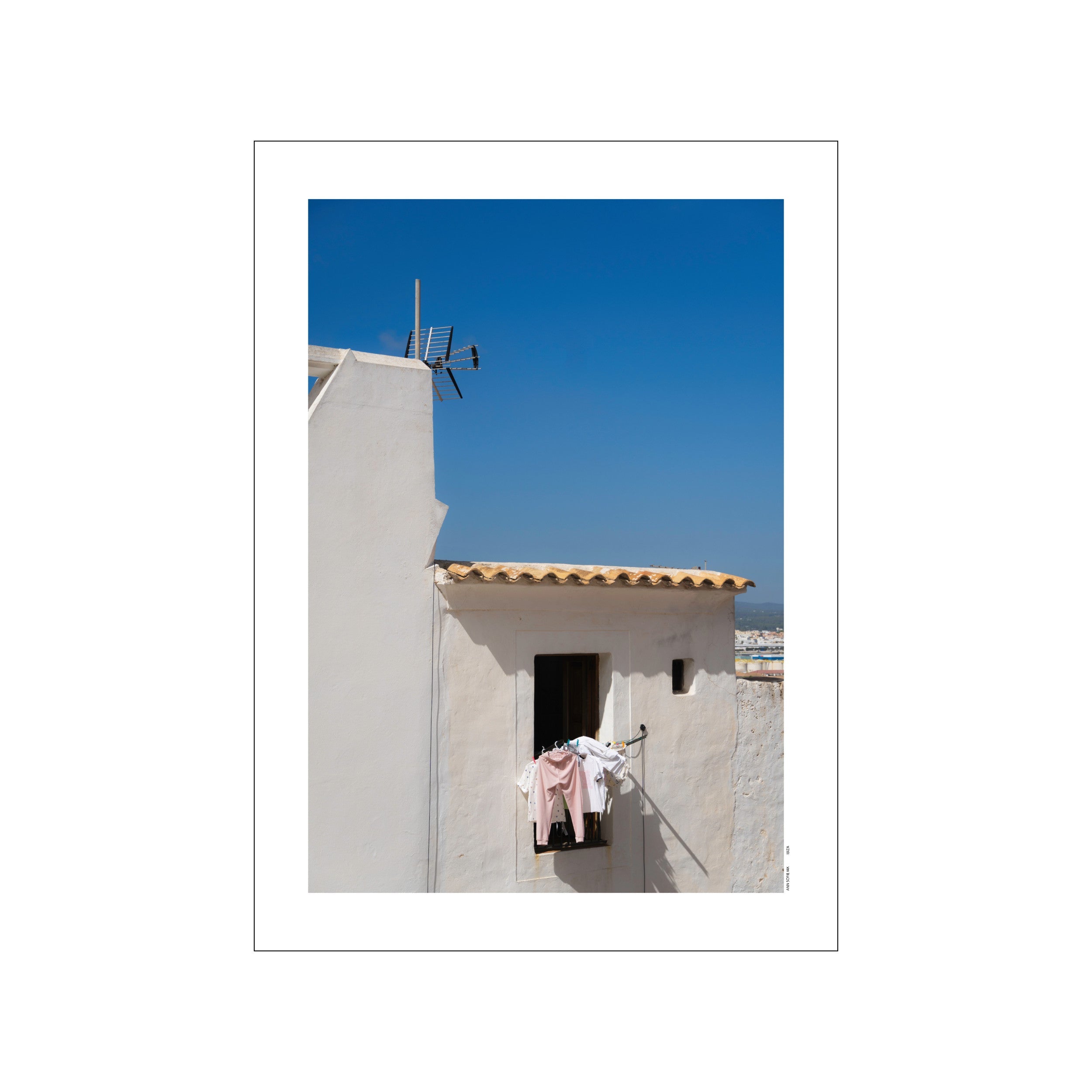 A bright photograph of a sun-drenched whitewashed building in Ibiza under a clear blue sky, with laundry hanging from a window, evoking a sense of peaceful island living.