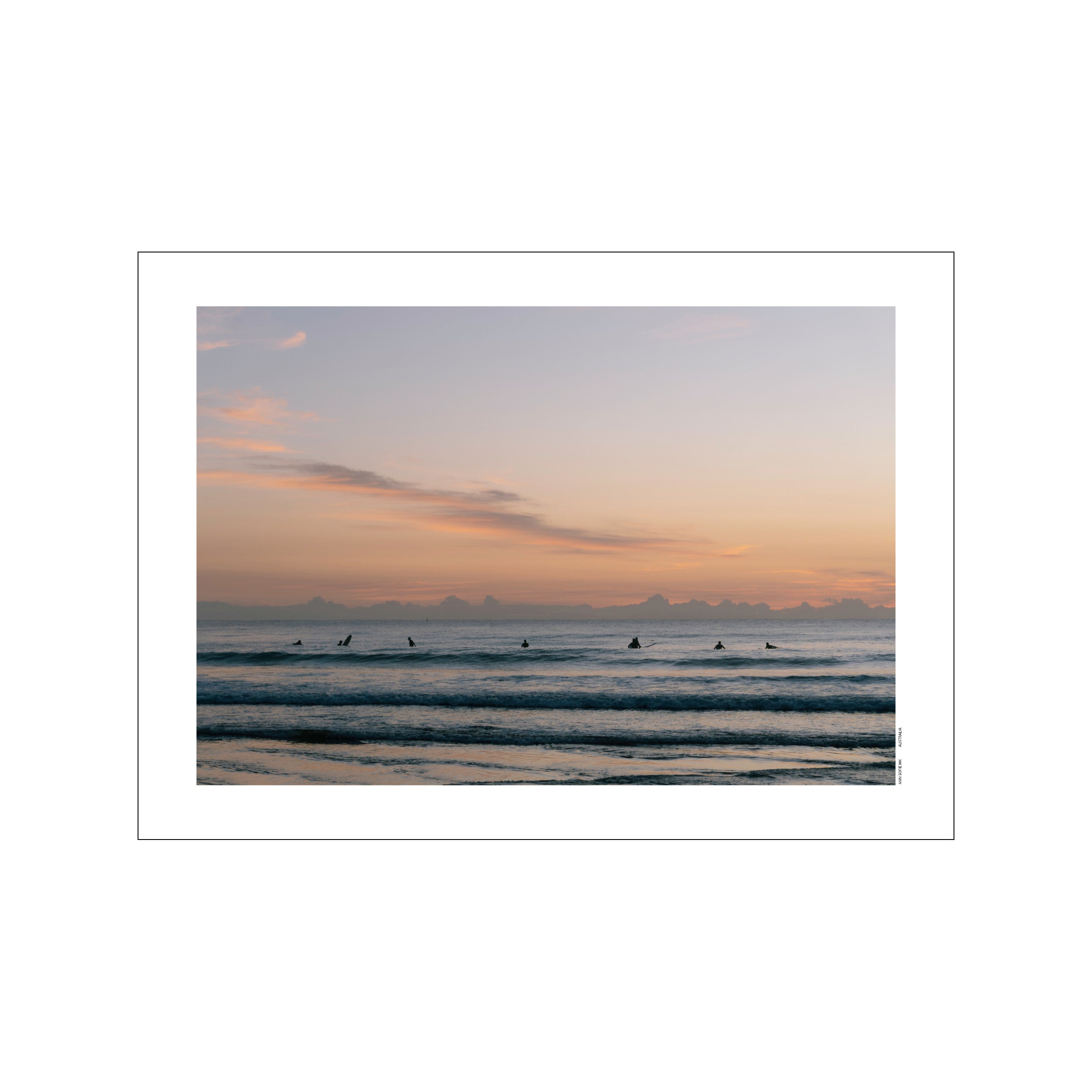 Surfers waiting on their boards in the calm ocean waves during a soft pink and lavender sunrise, evoking a serene and tranquil coastal atmosphere.