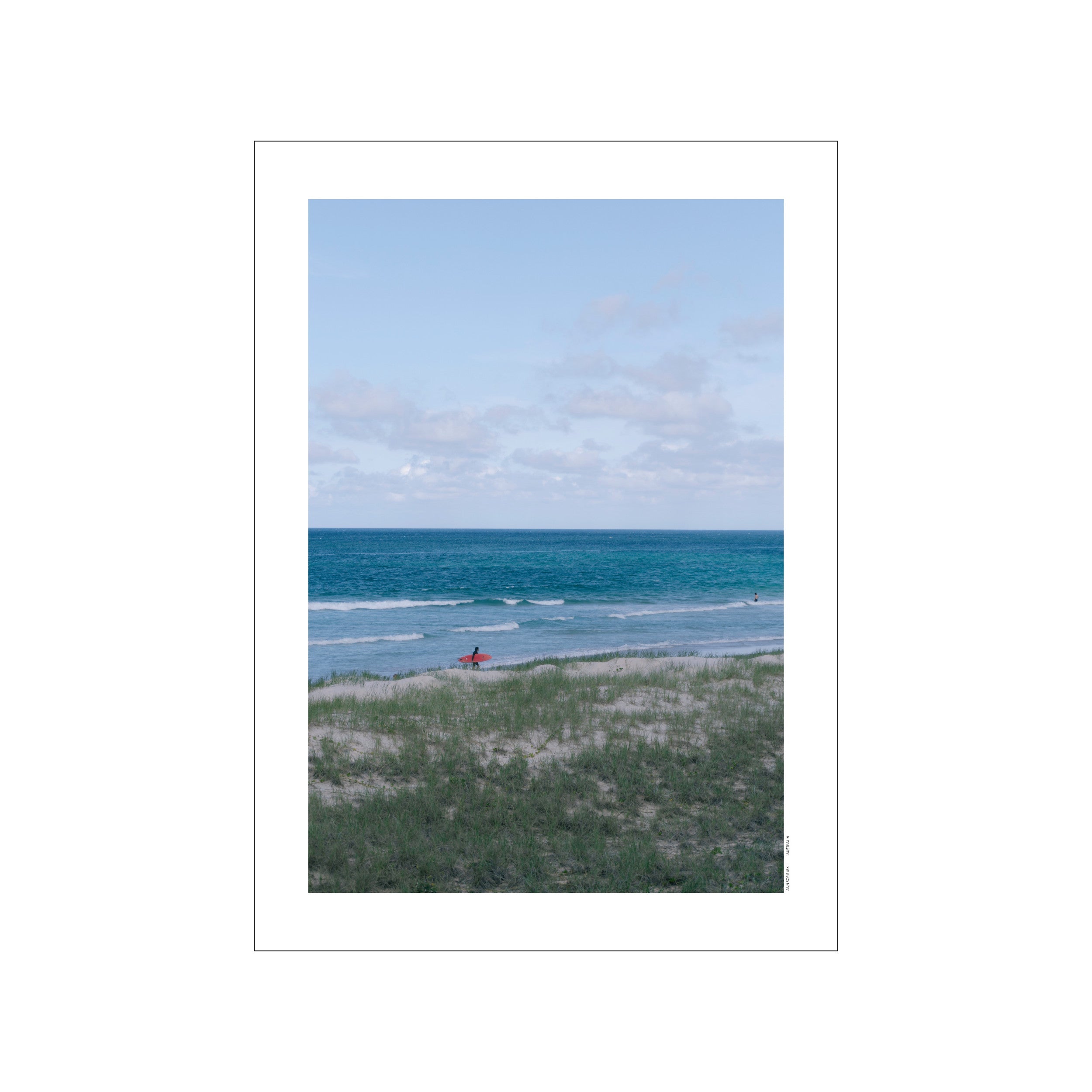 A person in silhouette walks along a sandy dune toward the turquoise ocean holding a bright red surfboard, under a pale blue sky with wispy clouds.