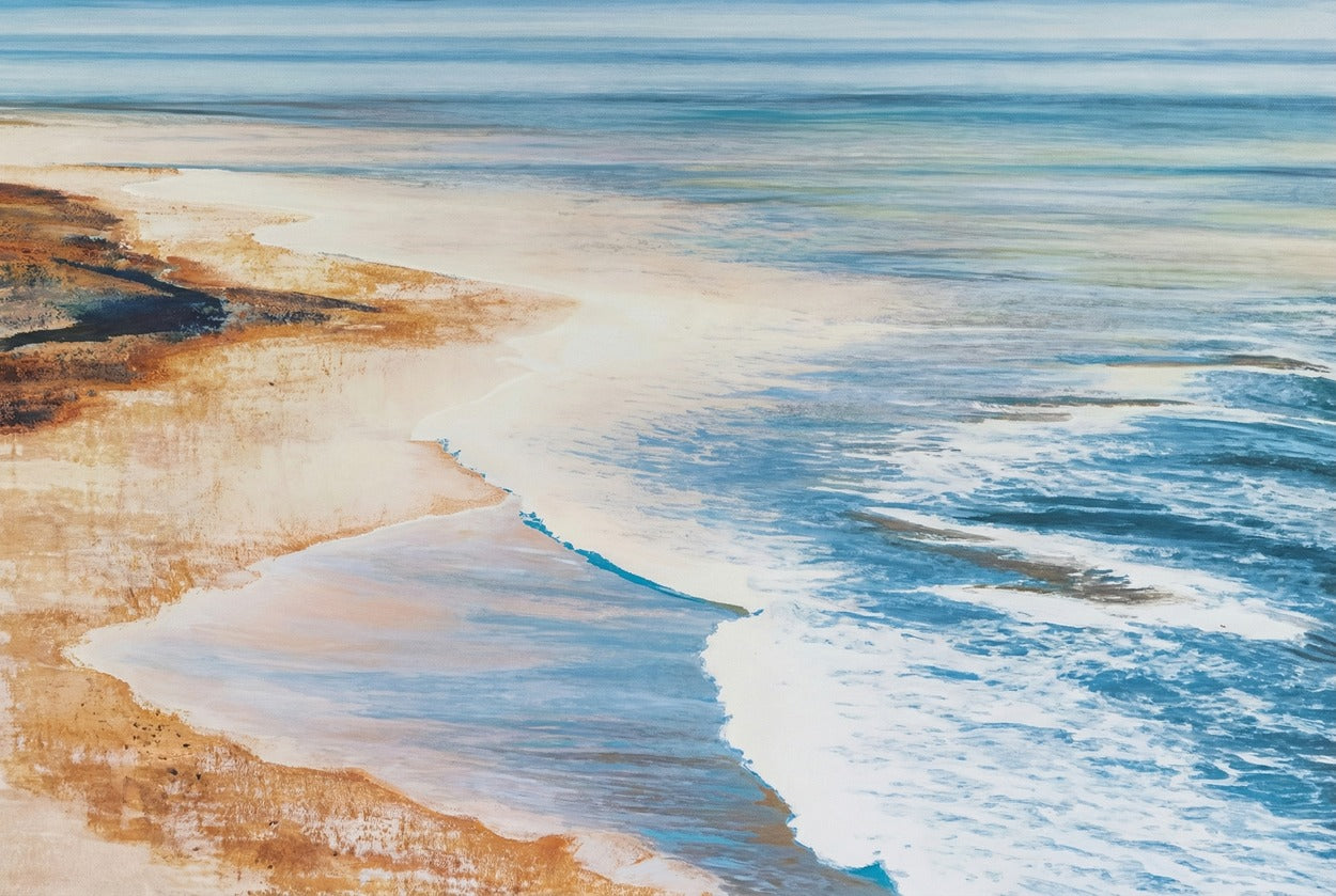 Blue surf curves along a warm sandy shoreline with rust-toned rock at the left edge