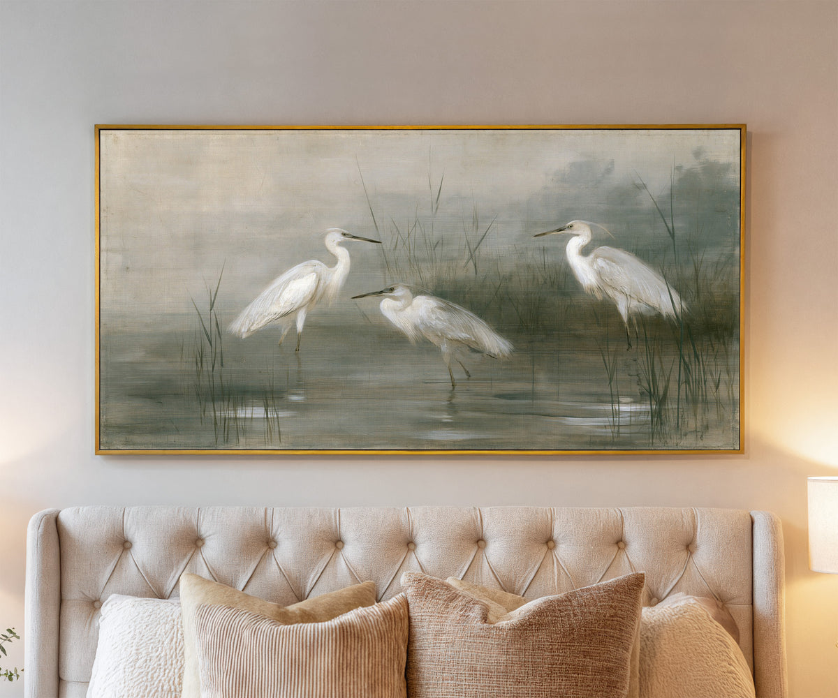 three white birds in shallow marsh water canvas wall art in a gold frame above a bed