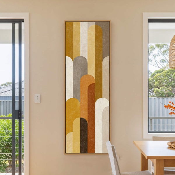 This vertical, abstract art print features a retro palette of ochre, brown, and cream, presented in a warm wood floater frame on a cream wall, with a window visible to.