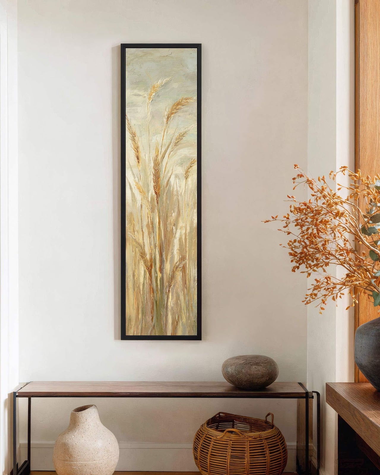 This vertical painting print showcases impressionistic golden wheat in an earthy palette, hung on a white hallway wall in a black floater frame above a wood console table.