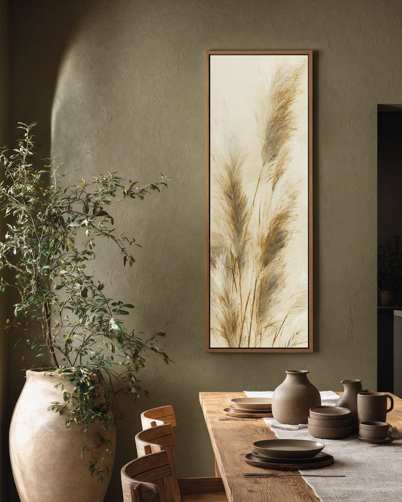 This vertical botanical art print of neutral pampas grass in a natural wood floater frame hangs on an olive green textured wall above a rustic wooden dining table.