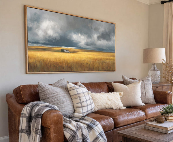 This horizontal canvas wall art features a landscape with a golden field under a dramatic, cloudy sky, and two houses in the distance, set against a light neutral wall with.