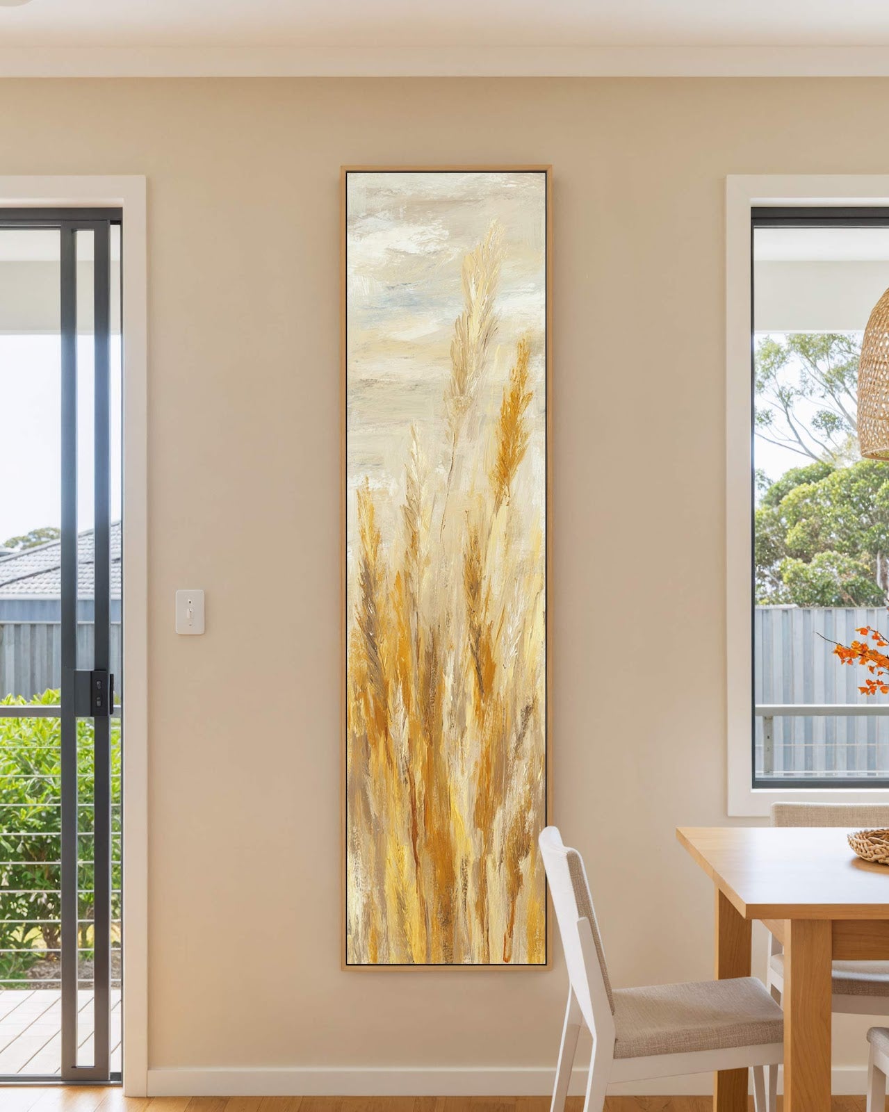 This vertical impressionist canvas print of golden wheat in an ochre palette hangs on a beige wall in a dining room with a wooden table and oak floater frame.