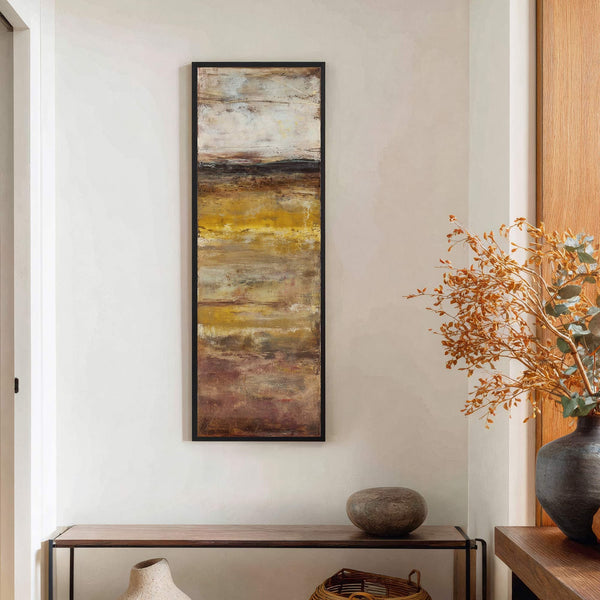 This vertical, abstract art print displays a warm palette of ochre, brown, and cream, with textured brushstrokes, framed in black and hanging on a textured cream wall above a wooden.
