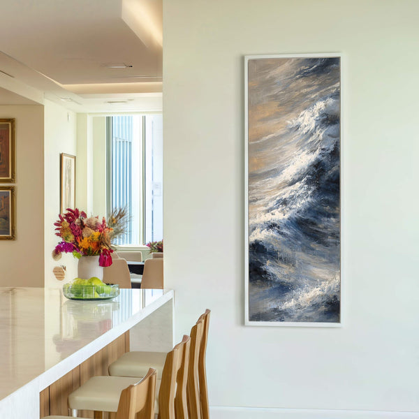 This vertical canvas wall art depicts a stormy wave with a palette of blues, grays, and sandy tones, displayed on a light-colored wall in a modern room with a bar.