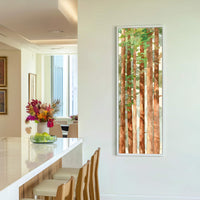 This vertical canvas print displays a watercolor painting of tall trees with a green and brown palette, framed in white and hung on a light-colored wall in a modern room.