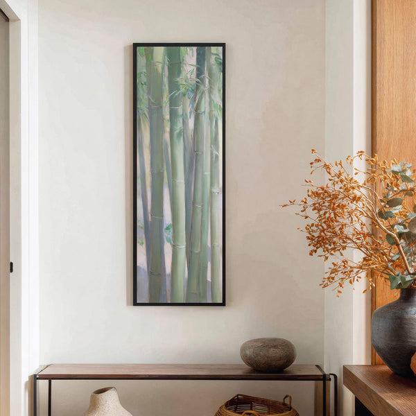 A vertical art print features a light green palette with painted bamboo stalks in a painterly style, framed in black and hung on a light-colored wall above a console table.