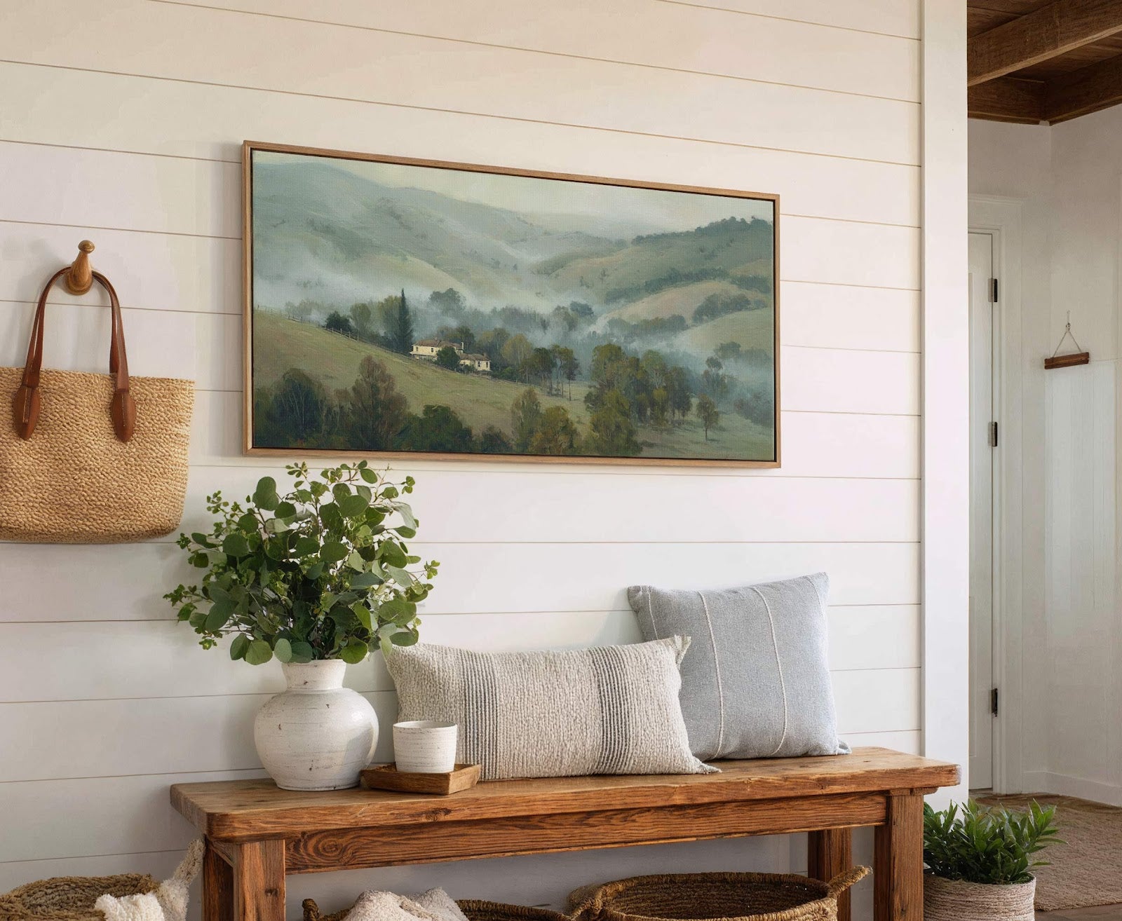 This canvas wall art features a misty landscape painting with green hills and houses in a soft palette, displayed horizontally in a natural wood frame on a white shiplap wall.