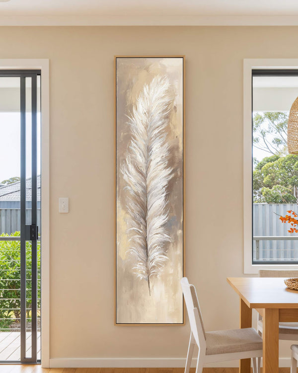 A vertical canvas print features an impressionist white feather in a neutral palette, displayed in an oak floater frame on a beige dining room wall beside a wooden table.