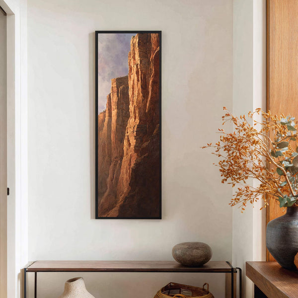 This vertical canvas print features a dramatic cliff face with warm, earthy tones and a cloudy sky, framed in black and displayed above a console table on a neutral wall.