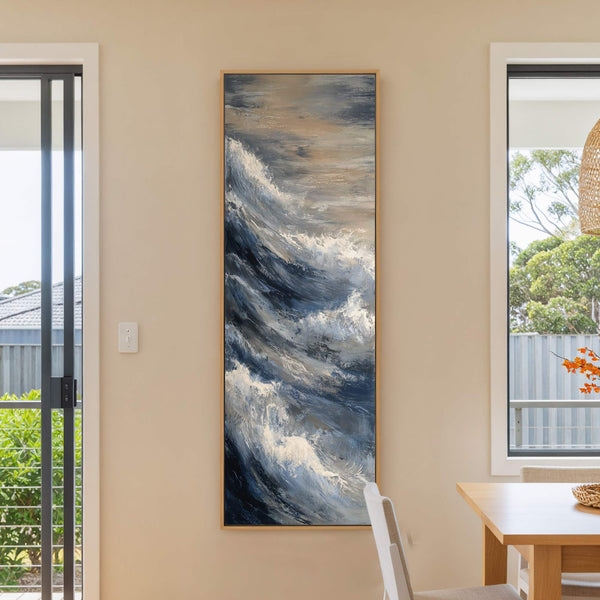 This vertical canvas wall art features an abstract ocean wave with a palette of blues, grays, and whites, framed in natural wood, hanging on a beige wall next to a.