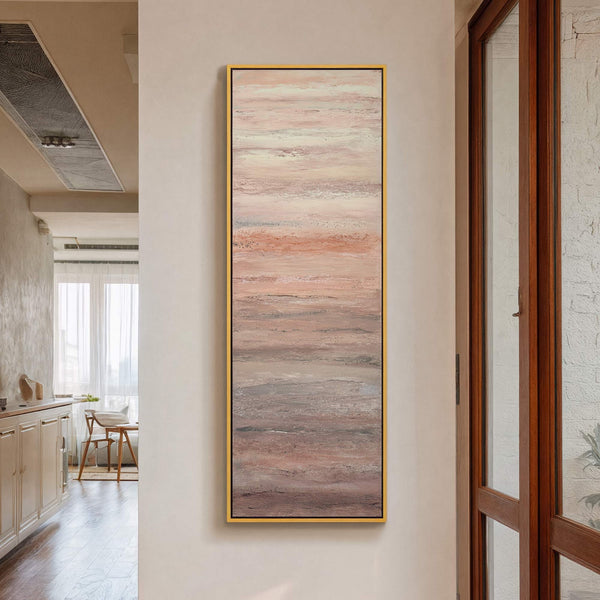 This vertical canvas wall art features an abstract landscape in a soft palette of blush pinks, grays, and creams, framed by a thin gold floater frame, displayed on a cream-colored.
