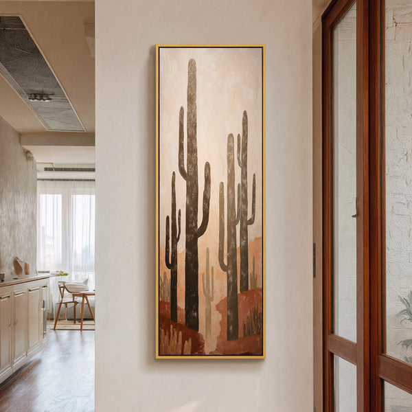This vertical canvas print features a stylized desert landscape with saguaro cacti in earthy tones, framed in gold and hanging on a neutral wall, with a wooden door visible to.