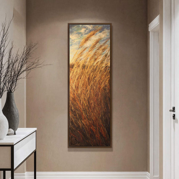 This vertical canvas wall art depicts tall, golden grasses in an impressionistic style with a palette of warm earth tones and sky blue. It hangs on a taupe wall above.