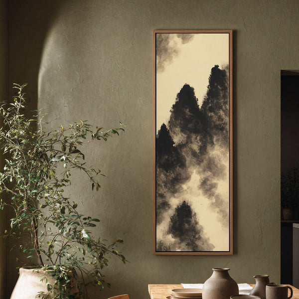This vertical art print of black and cream ink wash mountains hangs in a natural wood floater frame on a textured olive wall in a dining room beside a plant.