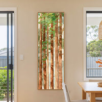 This vertical canvas wall art features a watercolor painting of redwood trees with a palette of greens and browns, framed in natural wood, hanging on a neutral wall in a.