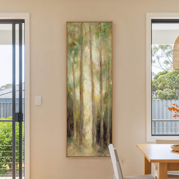 A vertical, Impressionist-style canvas wall art print depicts a sunlit forest with a palette of greens and yellows, framed in natural wood, hanging on a beige wall in a dining.