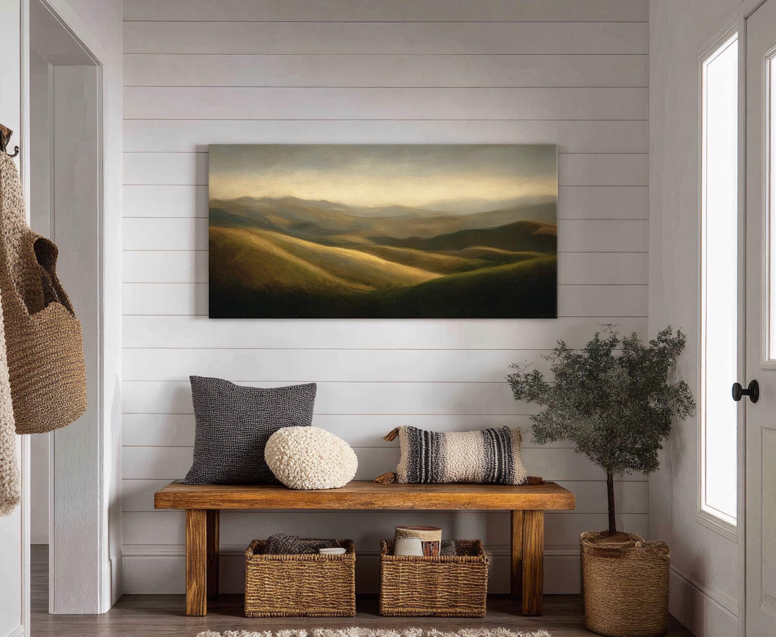 A horizontal canvas print depicts rolling hills bathed in golden light, with a muted palette and impressionistic style, displayed above a wooden bench with pillows and baskets in a room.