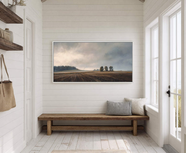 This horizontal canvas wall art depicts a serene landscape with muted earth tones and a dramatic sky, framed in white, hanging above a wooden bench in a room with white.