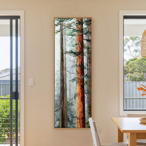 This vertical watercolor painting print depicts towering redwood trees in a misty forest, framed in natural wood, hanging on a pale peach wall above a wooden table.