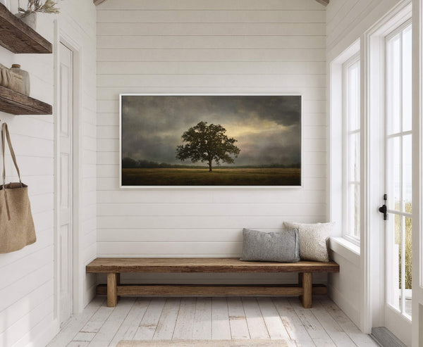 A horizontal canvas print of a lone tree in a field under a dramatic cloudy sky with a sliver of golden light, displayed above a rustic wooden bench with pillows.