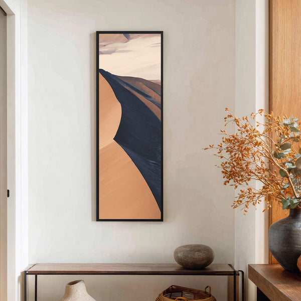 This vertical art print features a desert landscape in warm, muted tones with a black floater frame, displayed above a narrow wooden console table on a light, textured wall.