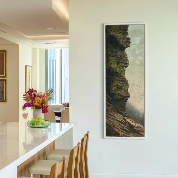 This vertical canvas wall art, with a naturalistic palette of greens and browns, depicts a rocky cliff face against a misty background, framed in white, hanging on a neutral wall.