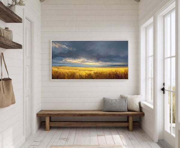 This horizontal canvas wall art features a landscape painting of a golden wheat field under dramatic, cloudy skies with a vibrant palette, displayed in a white frame above a rustic.