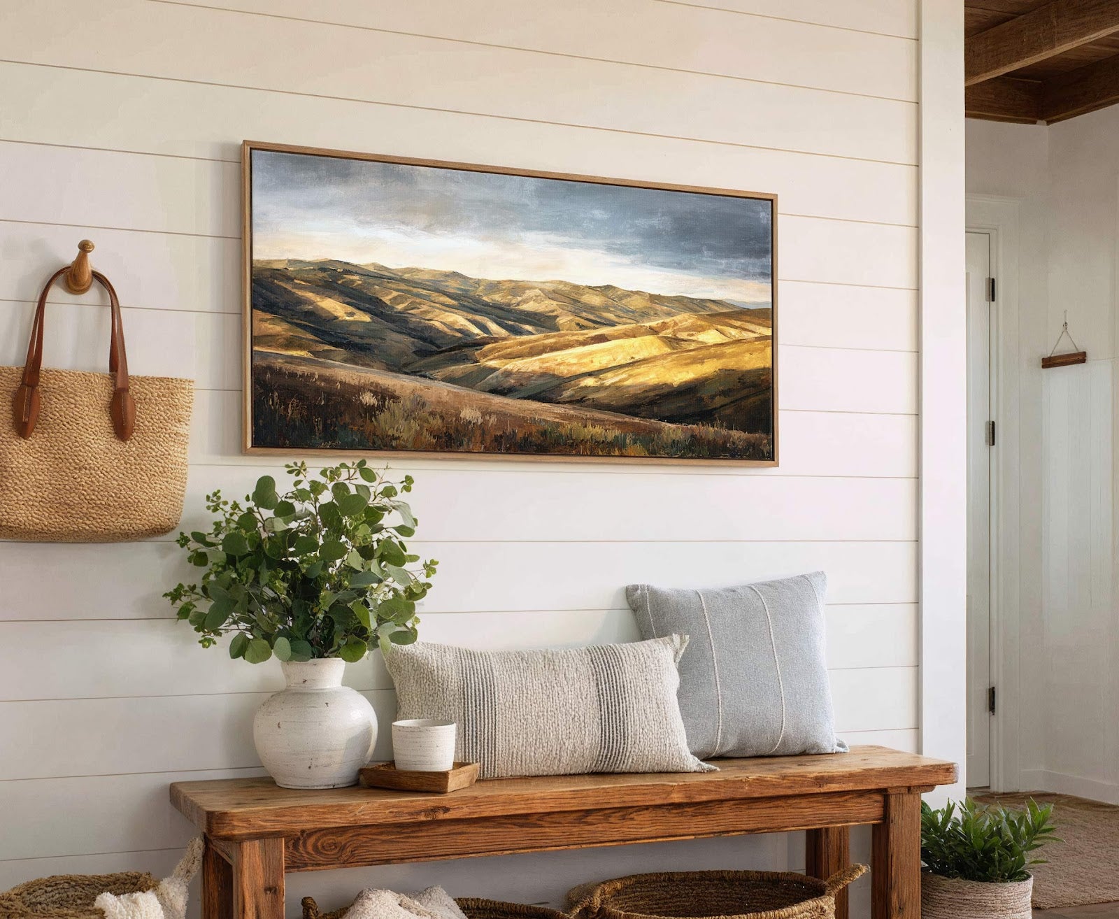 This horizontal canvas wall art depicts a warm, impressionistic landscape with golden hills under a dramatic sky, framed in natural wood, hanging above a wooden bench with decorative pillows and.